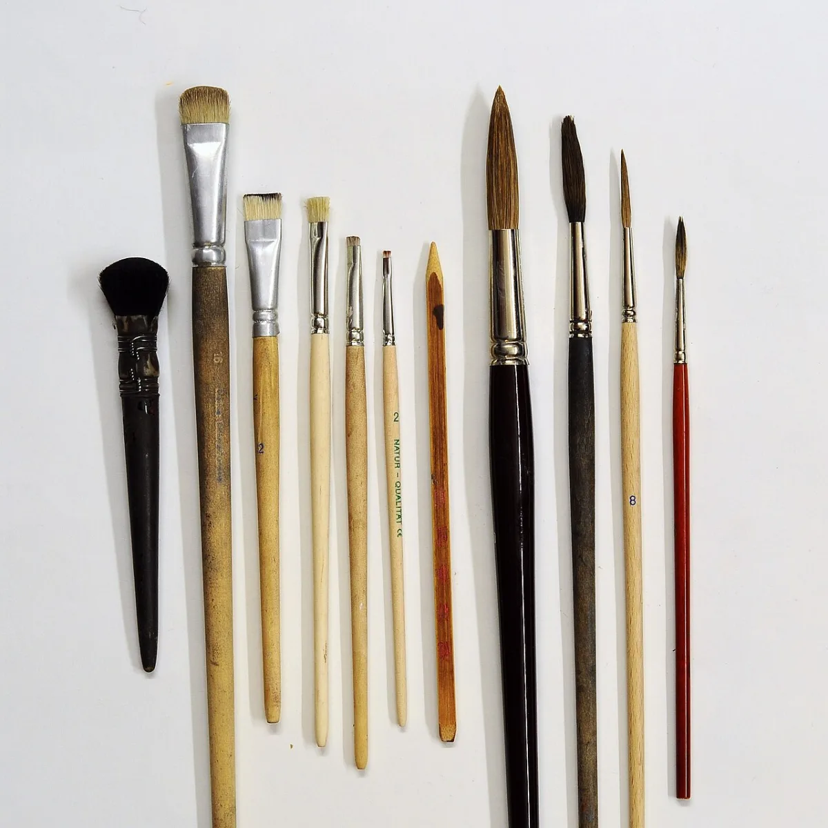 Een verzameling verschillende kwasten met houten handvatten en diverse haarpunten, naast elkaar op een witte ondergrond.